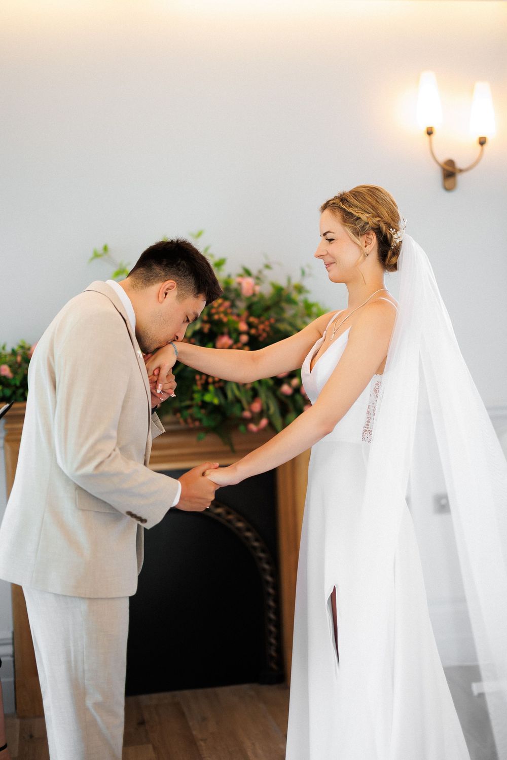 Groom in beige suit kissing bride's hand during wedding ceremony while bride smiles in white gown and veil.