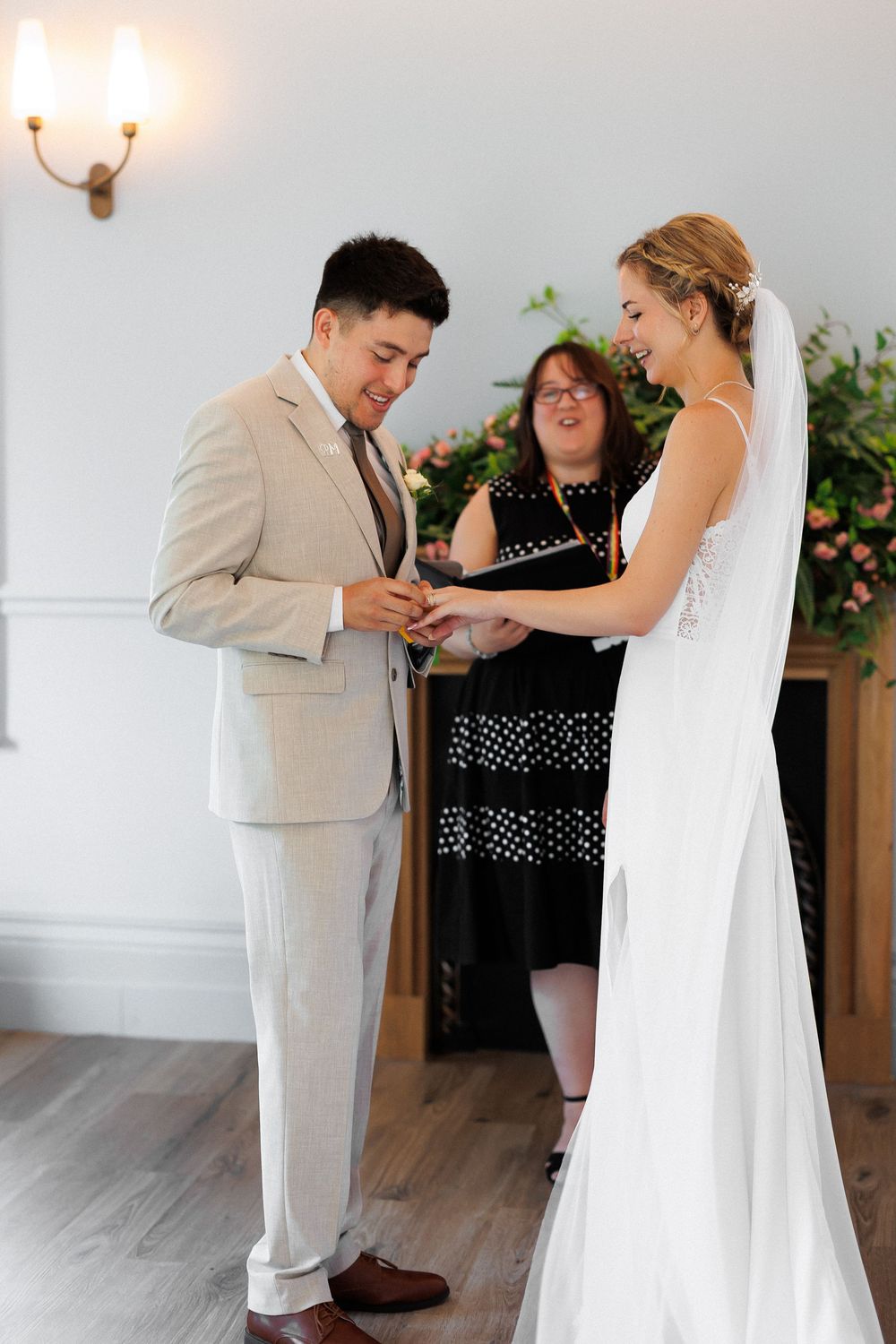 Groom placing wedding ring on bride's finger during indoor ceremony while officiant smiles in background.