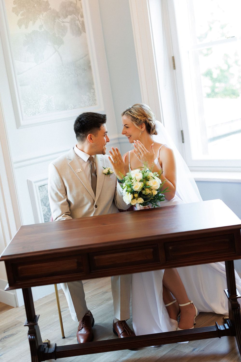 Bride and groom sitting at wooden desk, smiling at each other, bride holding white bouquet and showing wedding ring.