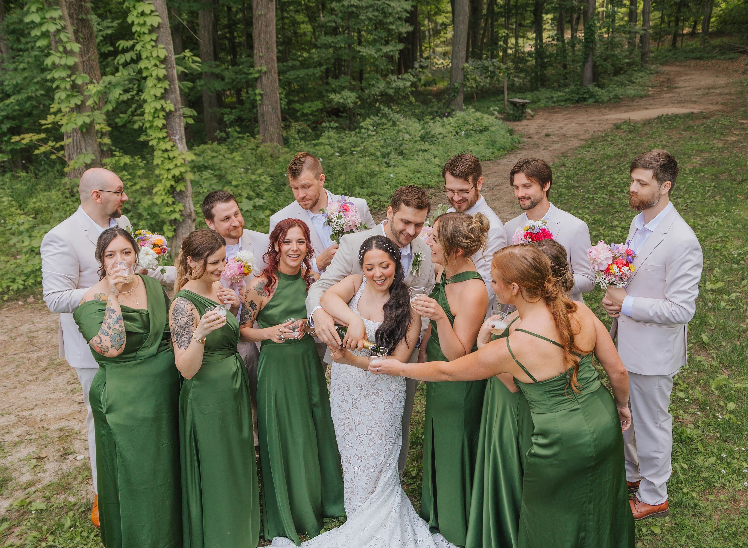 Wedding party in forest, bridesmaids in green gowns surround bride in white lace dress with groomsmen in light suits.