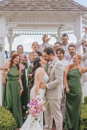 Bride and groom share a kiss under a white gazebo surrounded by cheering wedding party in green dresses and tan suits.