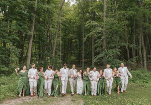 Wedding party walks through lush green forest, bridesmaids in sage green gowns and groomsmen in light gray suits.