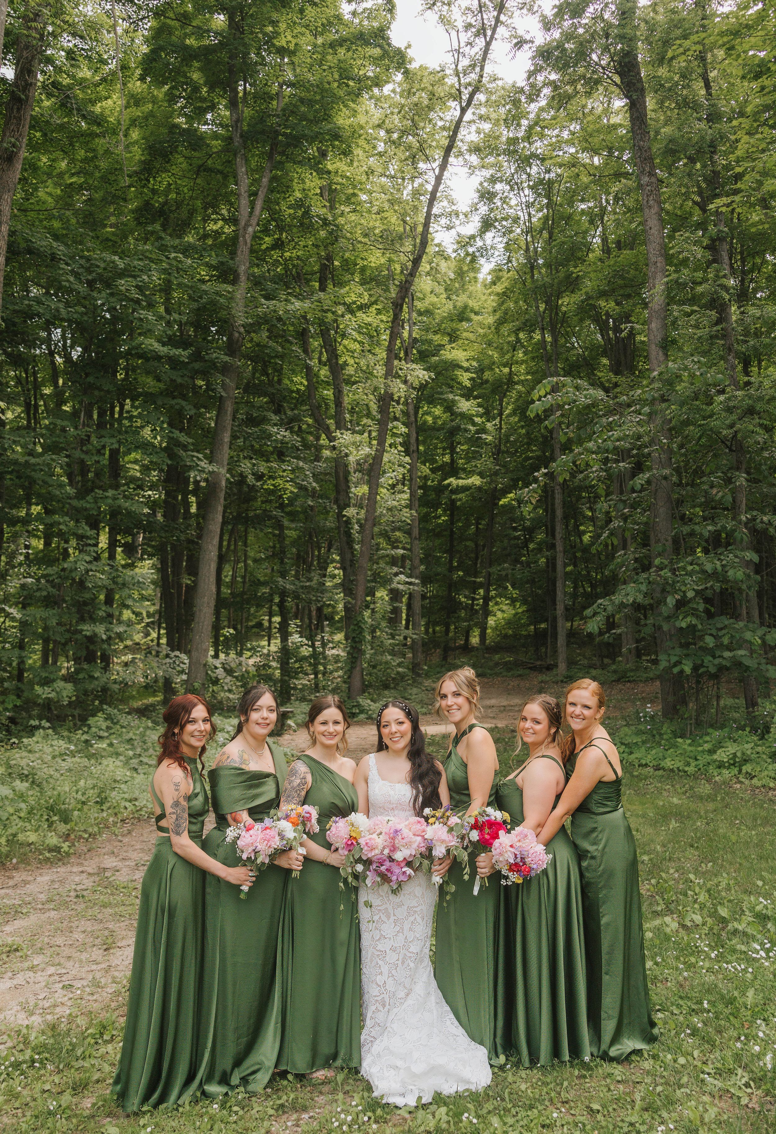 Bride in white lace gown with six bridesmaids in sage green dresses holding pink bouquets in a lush forest setting.