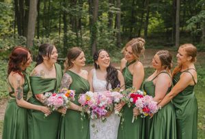 Bride in white lace gown laughing with six bridesmaids in green dresses holding colorful bouquets in a forest setting.