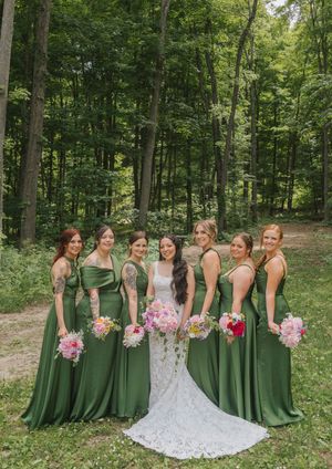 Bride in white lace gown with six bridesmaids in sage green dresses holding colorful bouquets in a forest setting.
