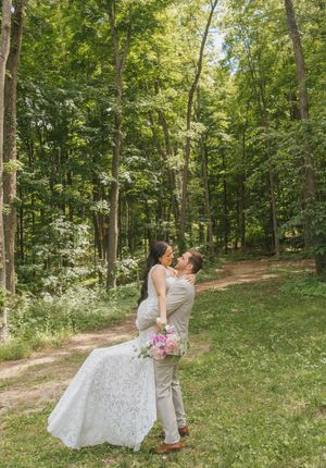 Bride and groom share a romantic embrace in a lush green forest clearing, bride holding a pink bouquet in wedding attire.