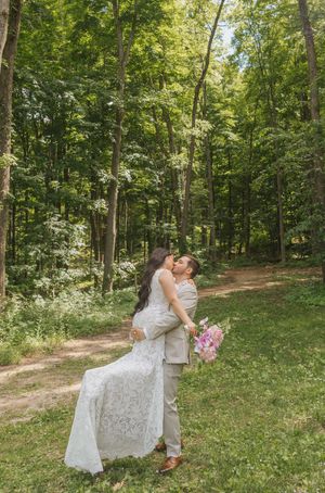 A couple shares a romantic kiss in a lush green forest, bride in white lace gown holding pink bouquet, groom in tan suit.