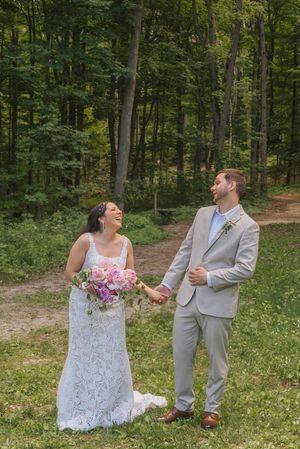 A laughing bride in lace gown holds bouquet while groom in gray suit holds her hand in a lush forest setting.