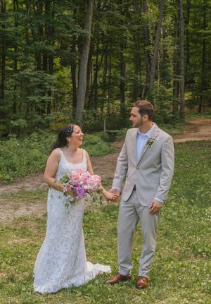 A smiling bride in lace gown holding pink bouquet holds hands with groom in tan suit outdoors in a lush green forest.