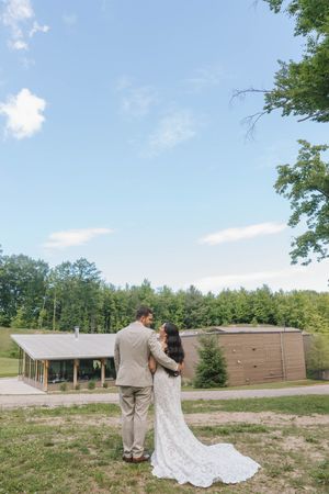 Bride in lace gown and groom in tan suit embrace outdoors near rustic building surrounded by lush green trees on wedding day.