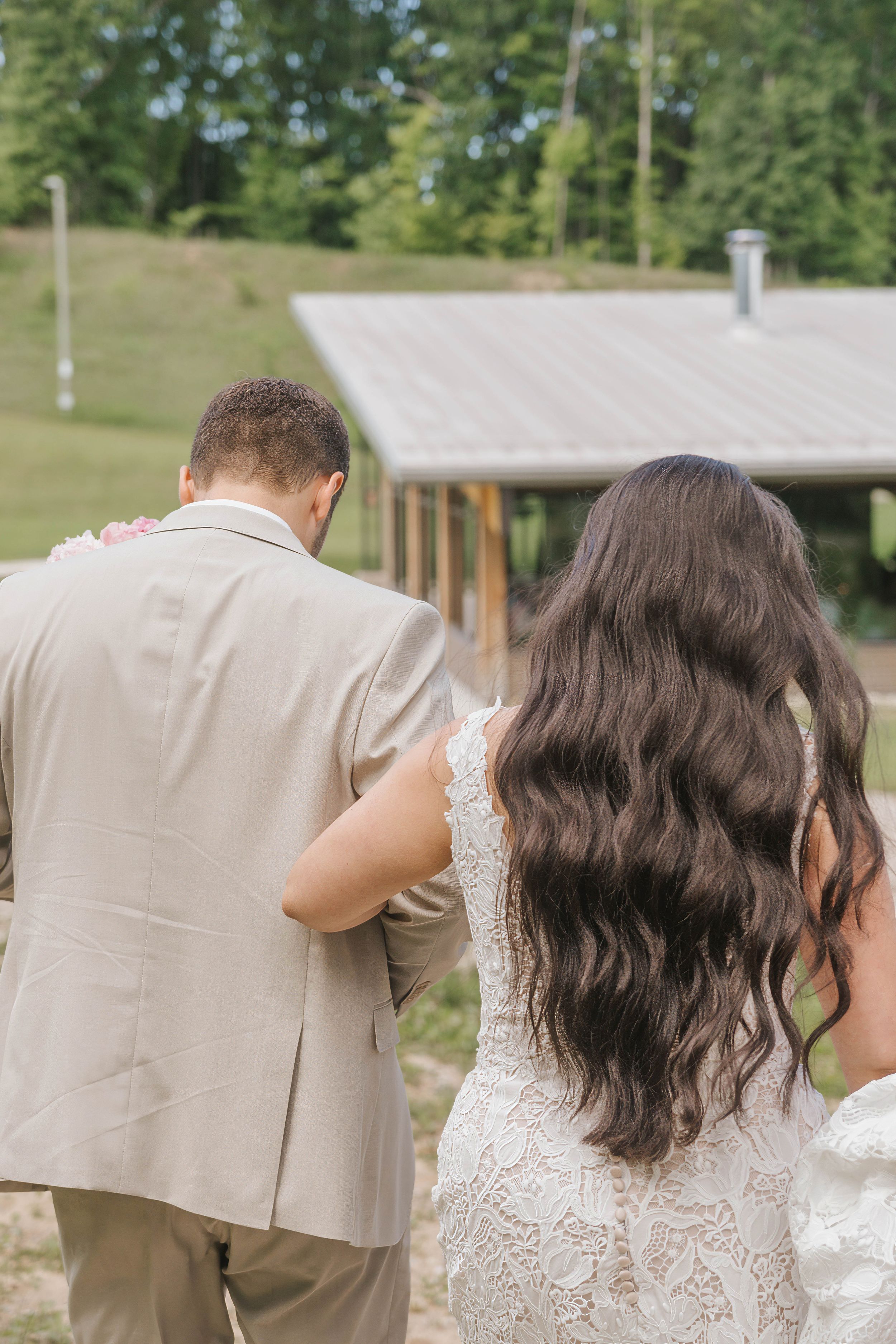 A bride in lace gown and groom in tan suit walk hand-in-hand toward a rustic cabin surrounded by lush greenery.