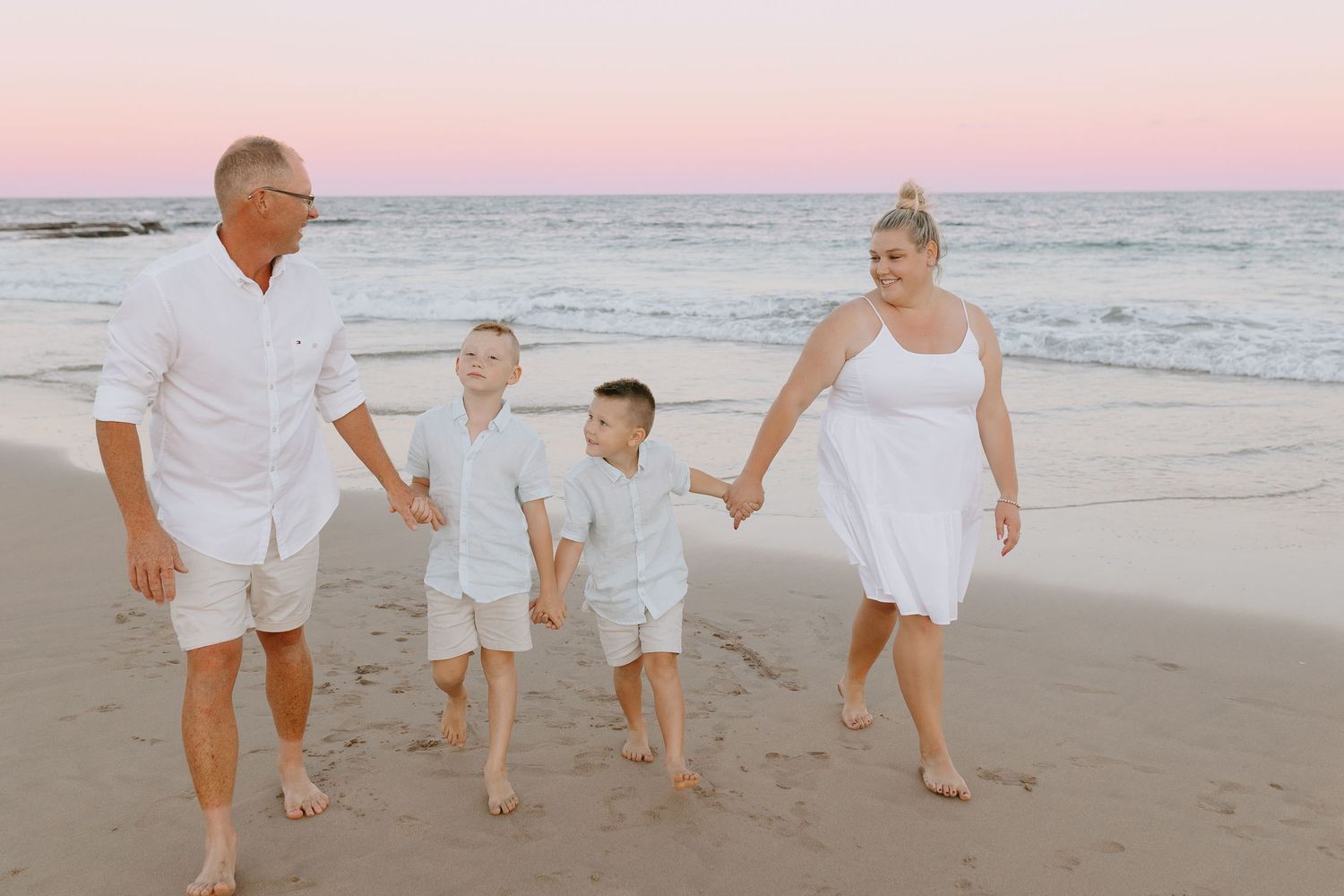 Family of four smiling together on rocky beach at golden sunset with ocean backdrop.