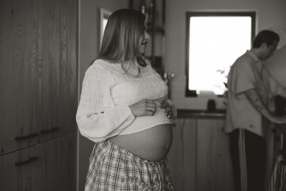 Black and white photo of a pregnant woman cradling her baby bump in a cozy home kitchen setting.