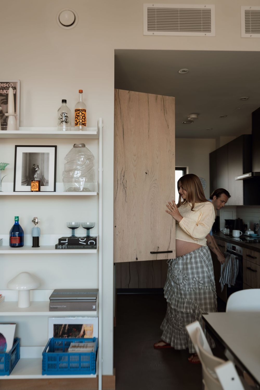 Pregnant woman in plaid skirt stands near modern kitchen cabinets while partner looks on, beside a white shelving unit.