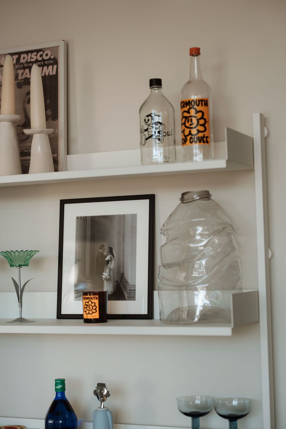 White wall shelves displaying glass bottles, framed black and white photo, candle holders, and decorative glassware in a modern home.