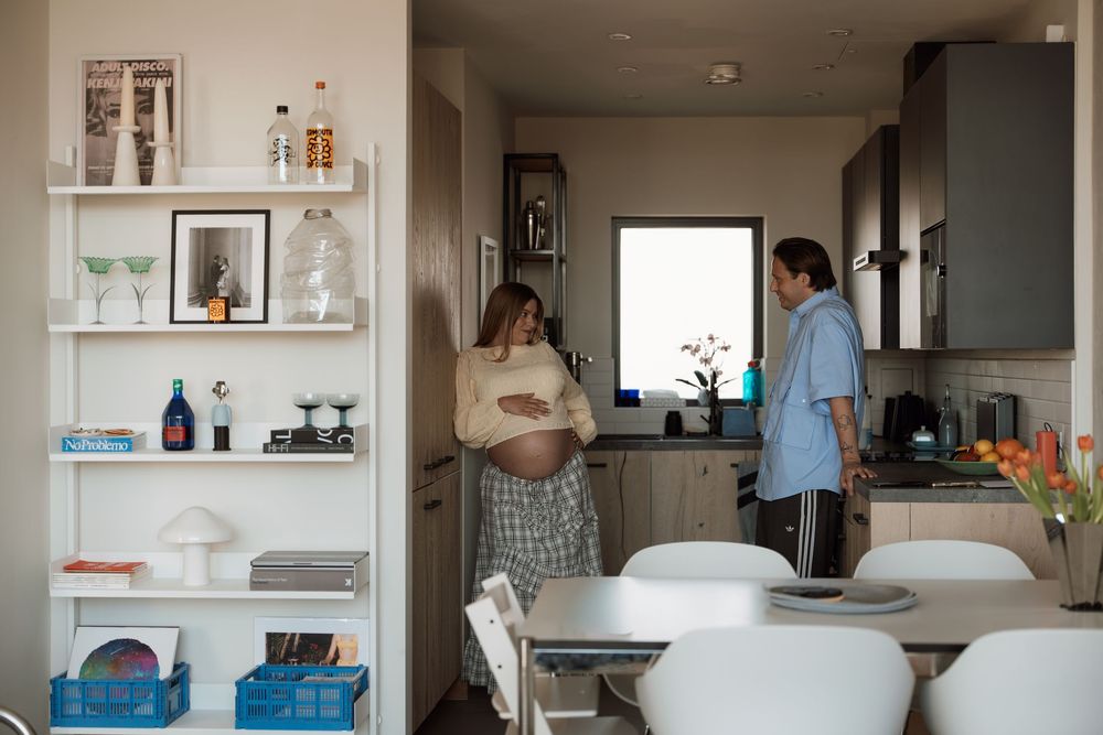 Pregnant woman and partner talking in a modern kitchen with white shelving unit displaying books and decorative items.