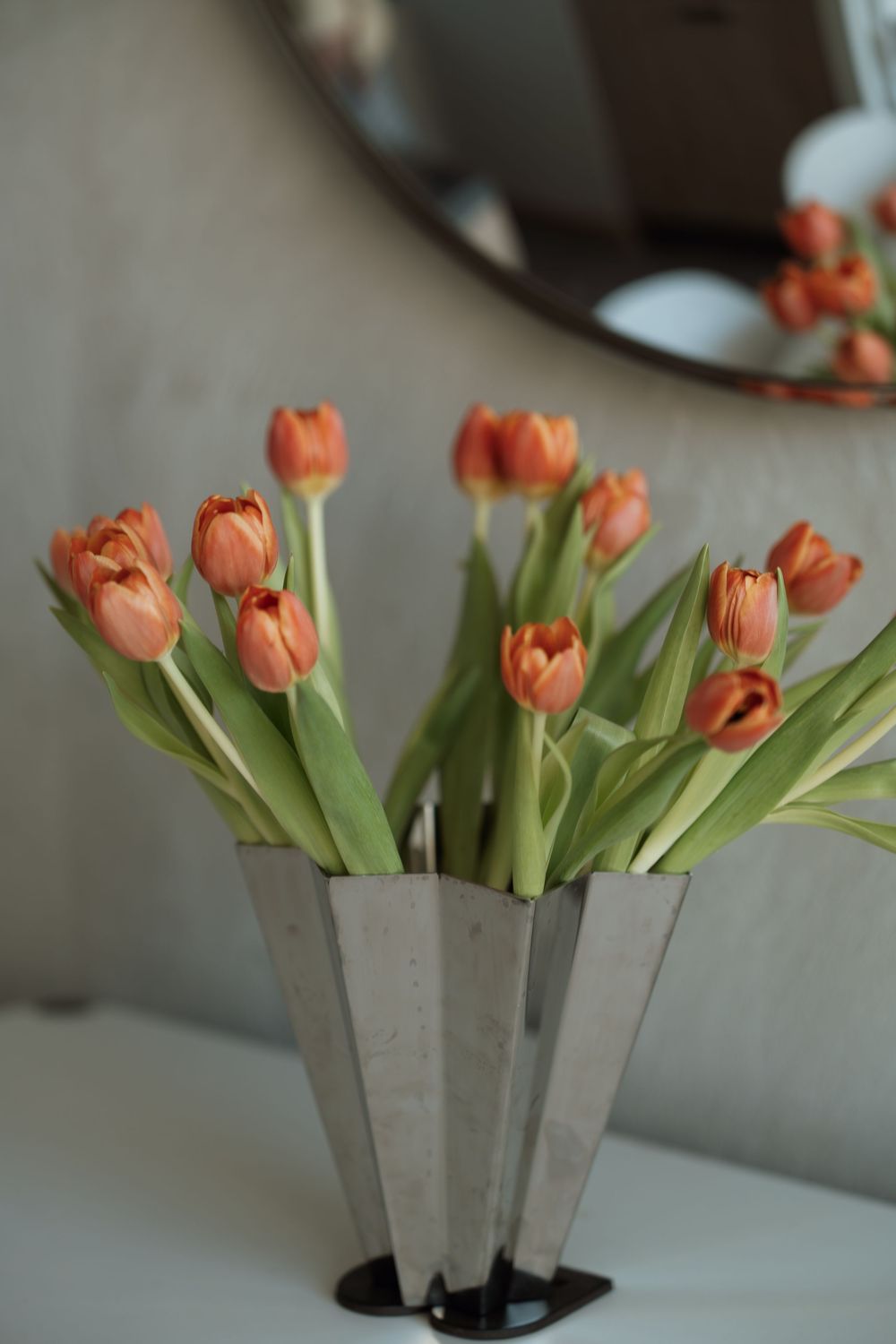 Orange tulips arranged in a modern silver geometric vase on a white surface with a round mirror in the background.