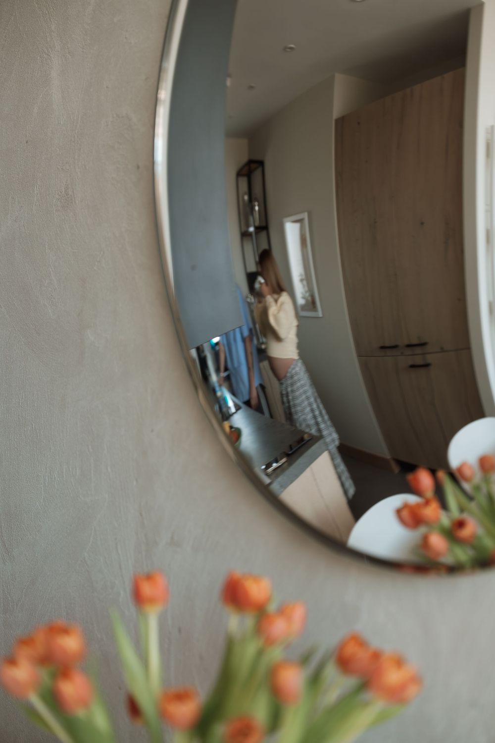 Round mirror on gray wall reflecting woman in kitchen, with orange tulips in foreground.