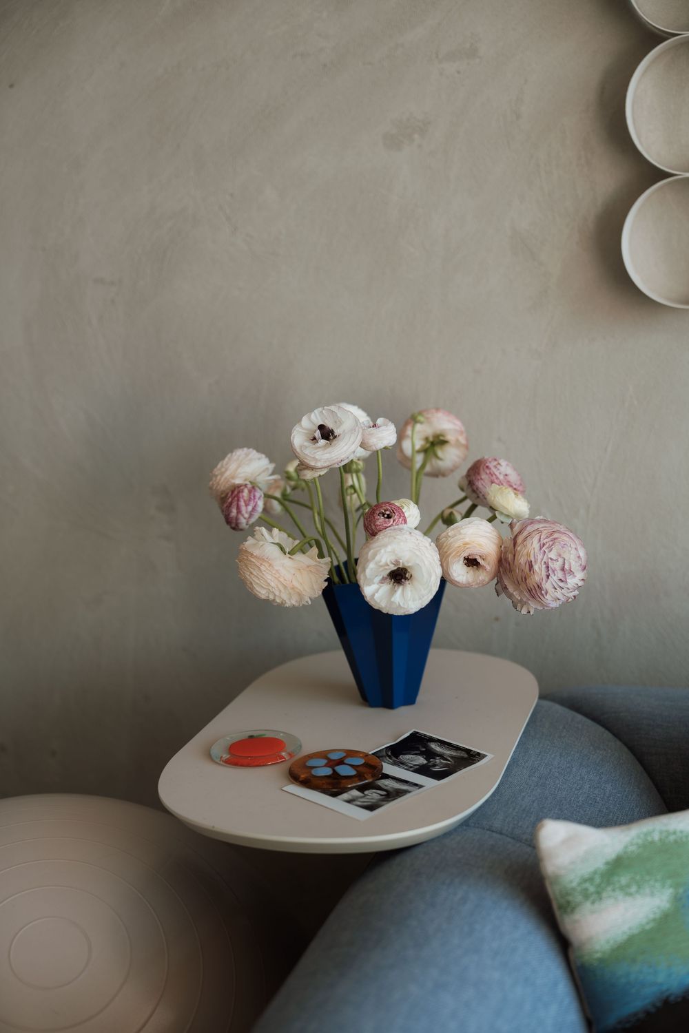 Blue vase with pink and white ranunculus flowers on a white side table beside a gray sofa in a modern interior.