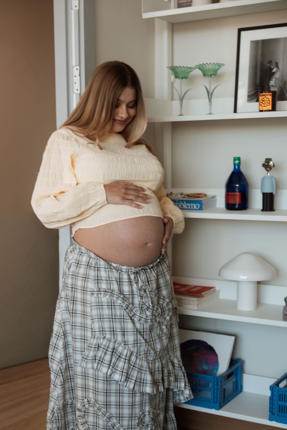 Pregnant woman in cream sweater and plaid skirt cradling her baby bump while standing near a white shelf.