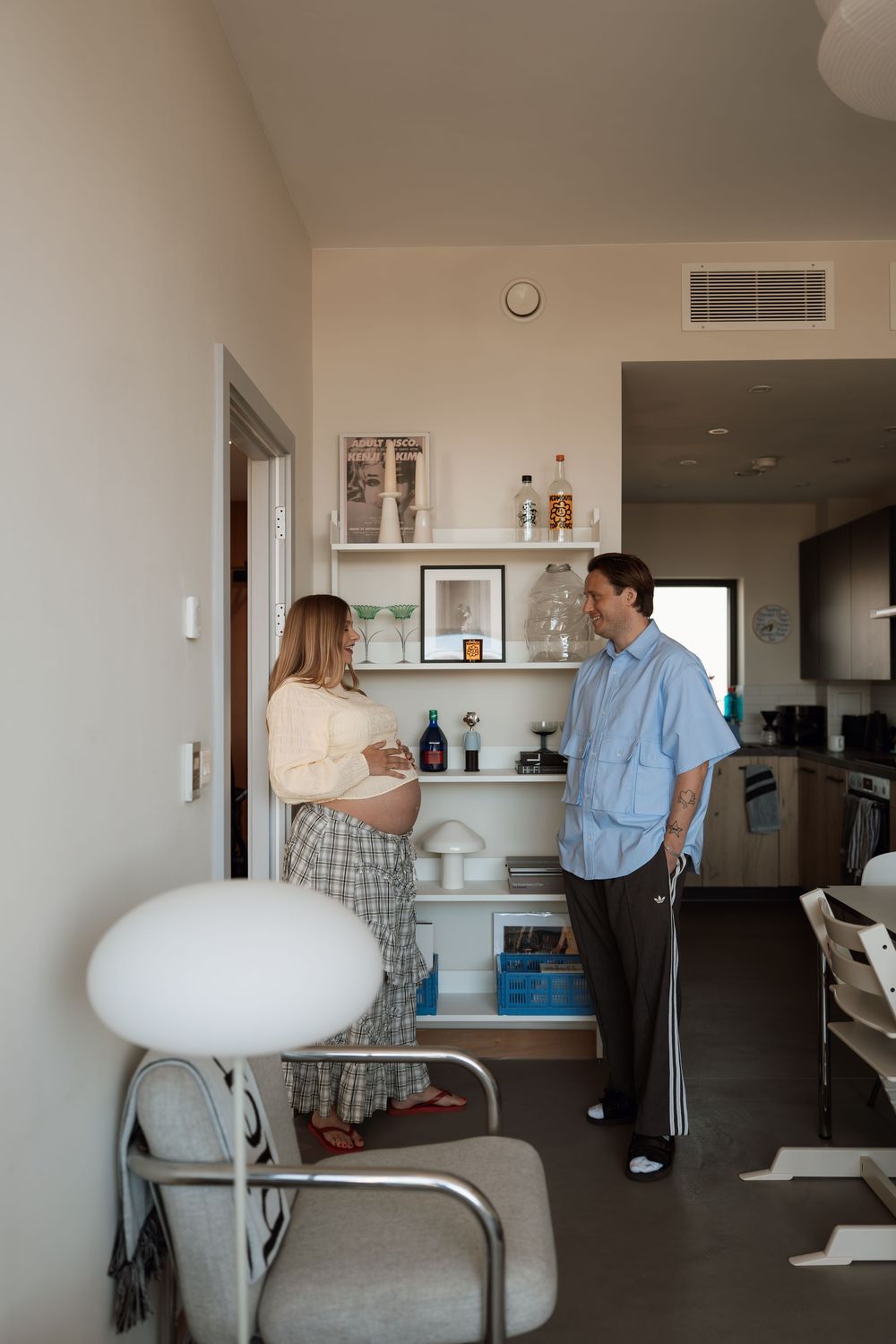 Pregnant woman and man conversing in a modern apartment living room with shelves and kitchen in background.