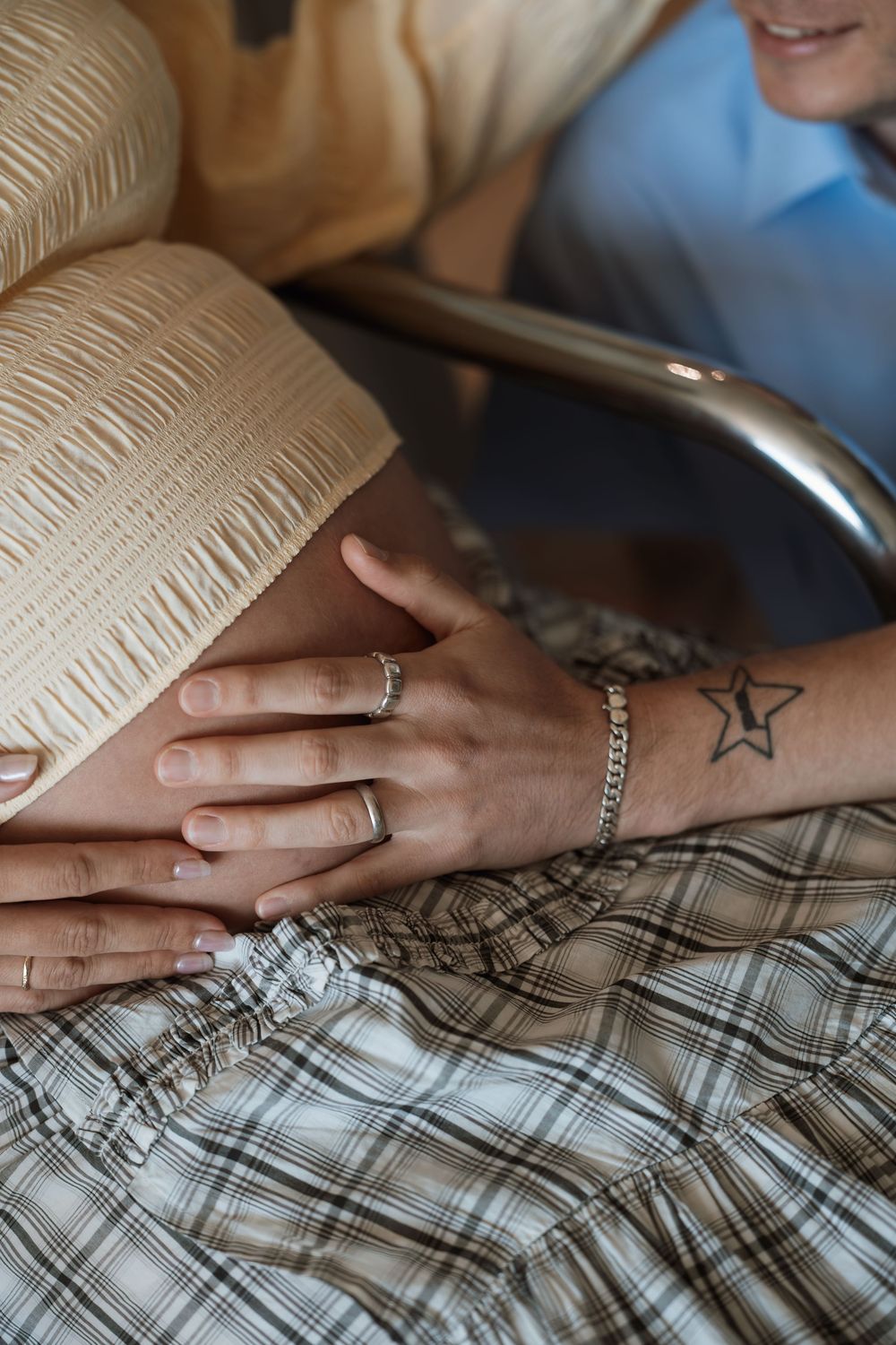 Close-up of hands with rings and star tattoo resting on plaid fabric near a hospital bed railing.