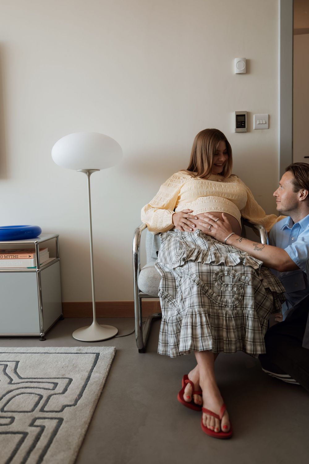 Pregnant woman in yellow sweater and plaid skirt sits while partner tenderly touches her baby bump in a cozy room.