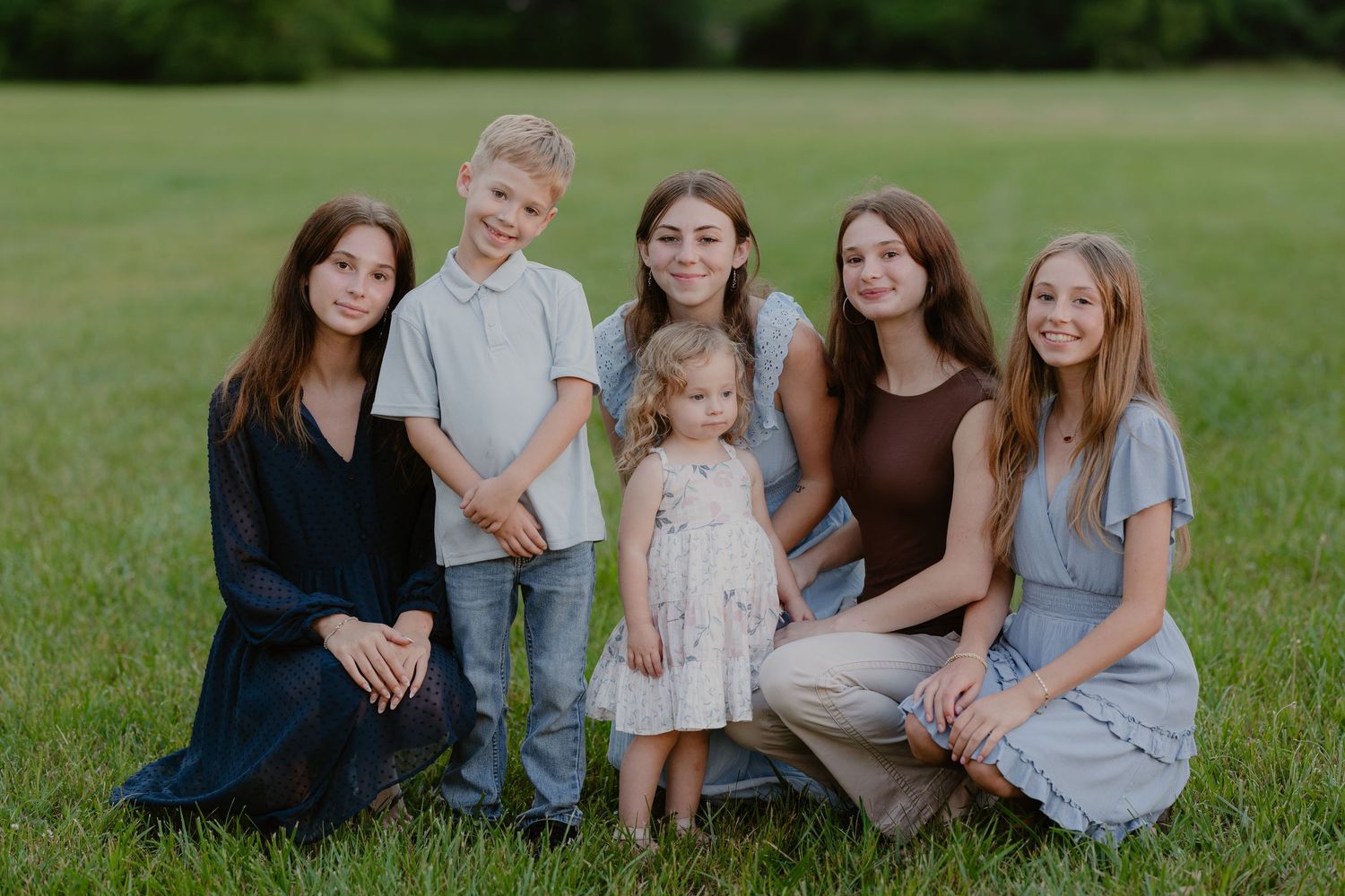 Family of six posing outdoors on green grass field, including four girls, one boy, and a toddler in white dress.
