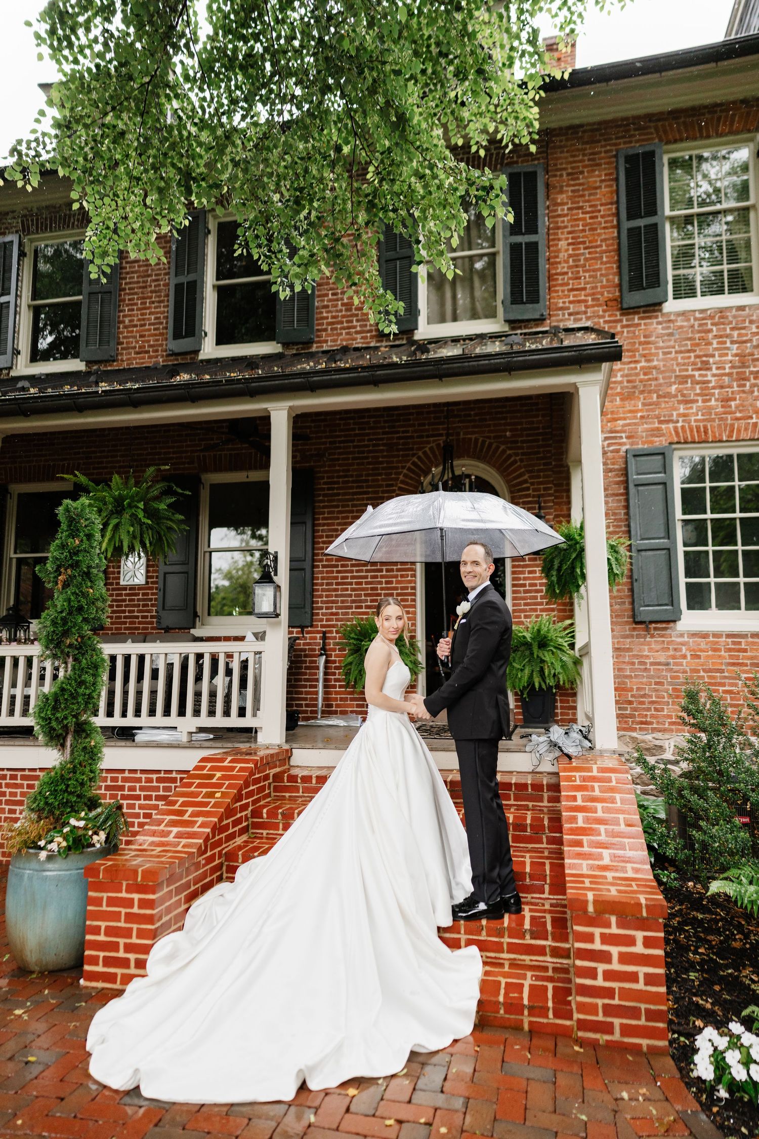 Bride and groom pose with a clear umbrella on brick steps outside a historic red brick colonial mansion.