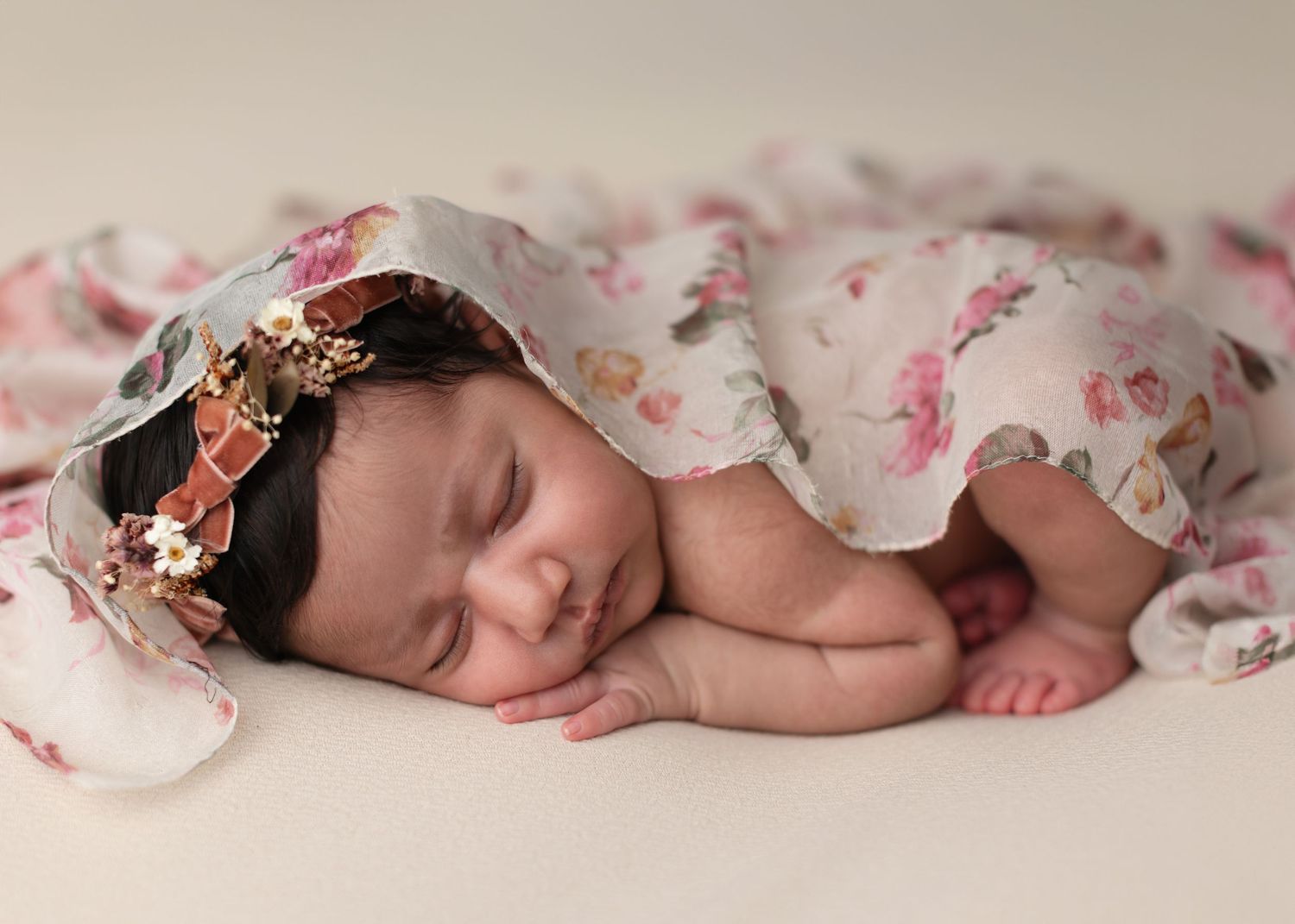 Sleeping newborn baby wrapped in a floral blanket with a flower headband on a beige background.