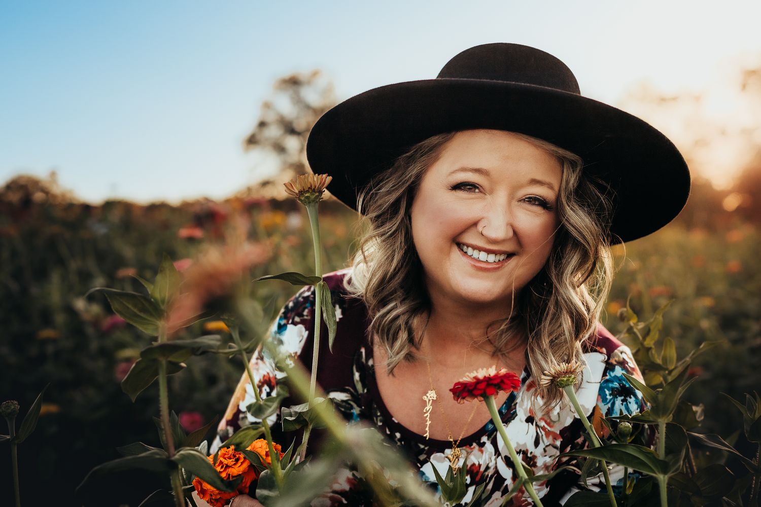 Smiling woman in black wide-brim hat holding colorful flowers in a sunlit field at golden hour.