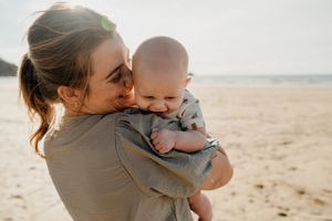 A young mother lovingly holds her smiling baby on a sunny beach, sharing a tender moment together.