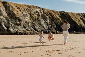 Family playing on sandy beach with rocky cliffs, parents and toddler enjoying sunny day by the ocean.