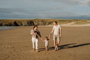Family of four walking on a sandy beach at golden hour, parents holding toddler's hands with cliffs in background.