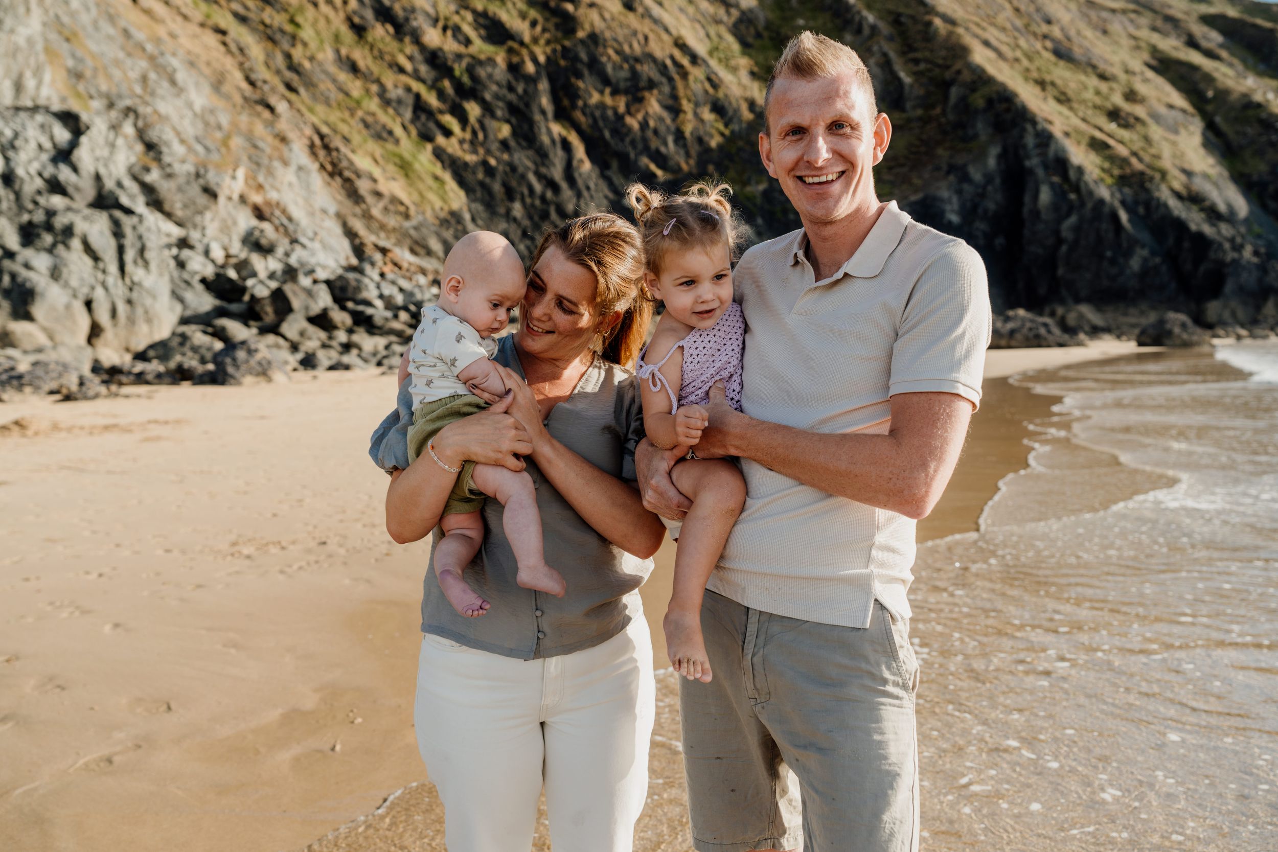 Happy family of four posing on a sandy beach, parents holding two young children with rocky cliffs in the background.