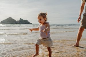 Toddler girl playing in ocean waves on a sandy beach with an adult nearby on a sunny day.