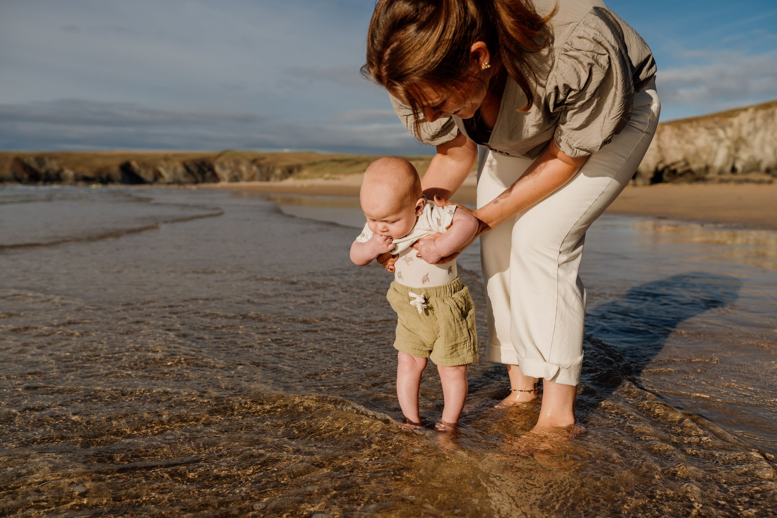 Mother helping baby stand in shallow ocean waves on a sandy beach during golden hour sunset.