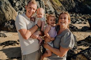 Happy family of four smiling on a rocky beach, parents holding two young children in casual summer outfits.