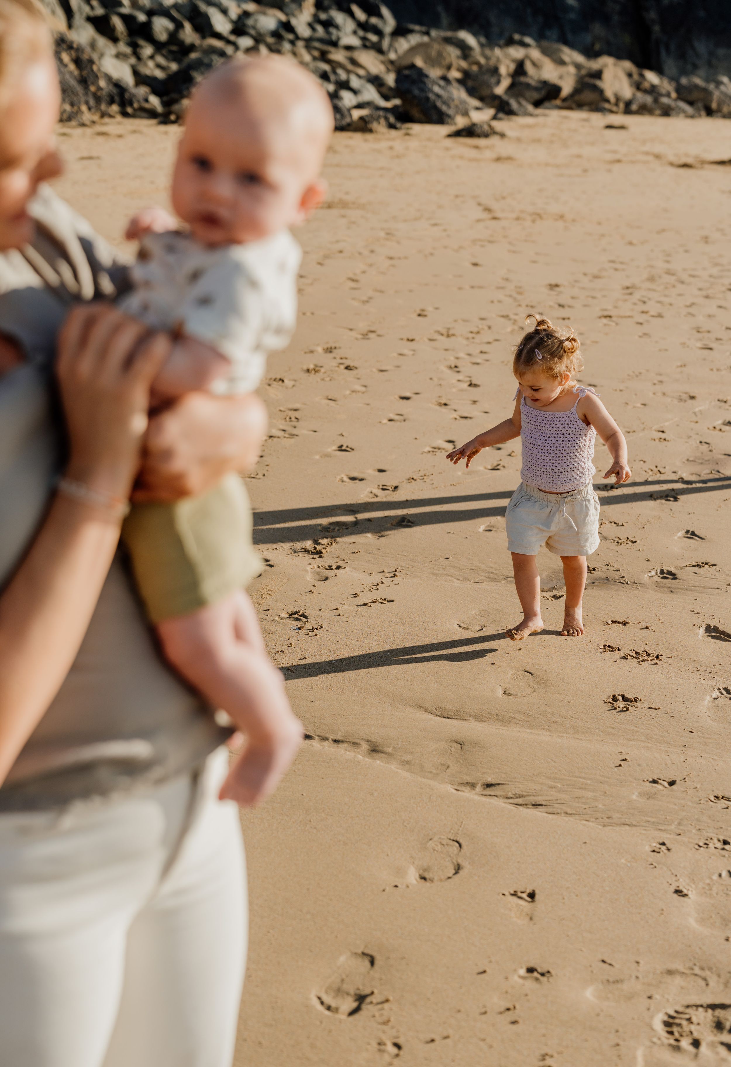 A mother holds her baby on a sunny beach while a toddler walks barefoot on the sand nearby.