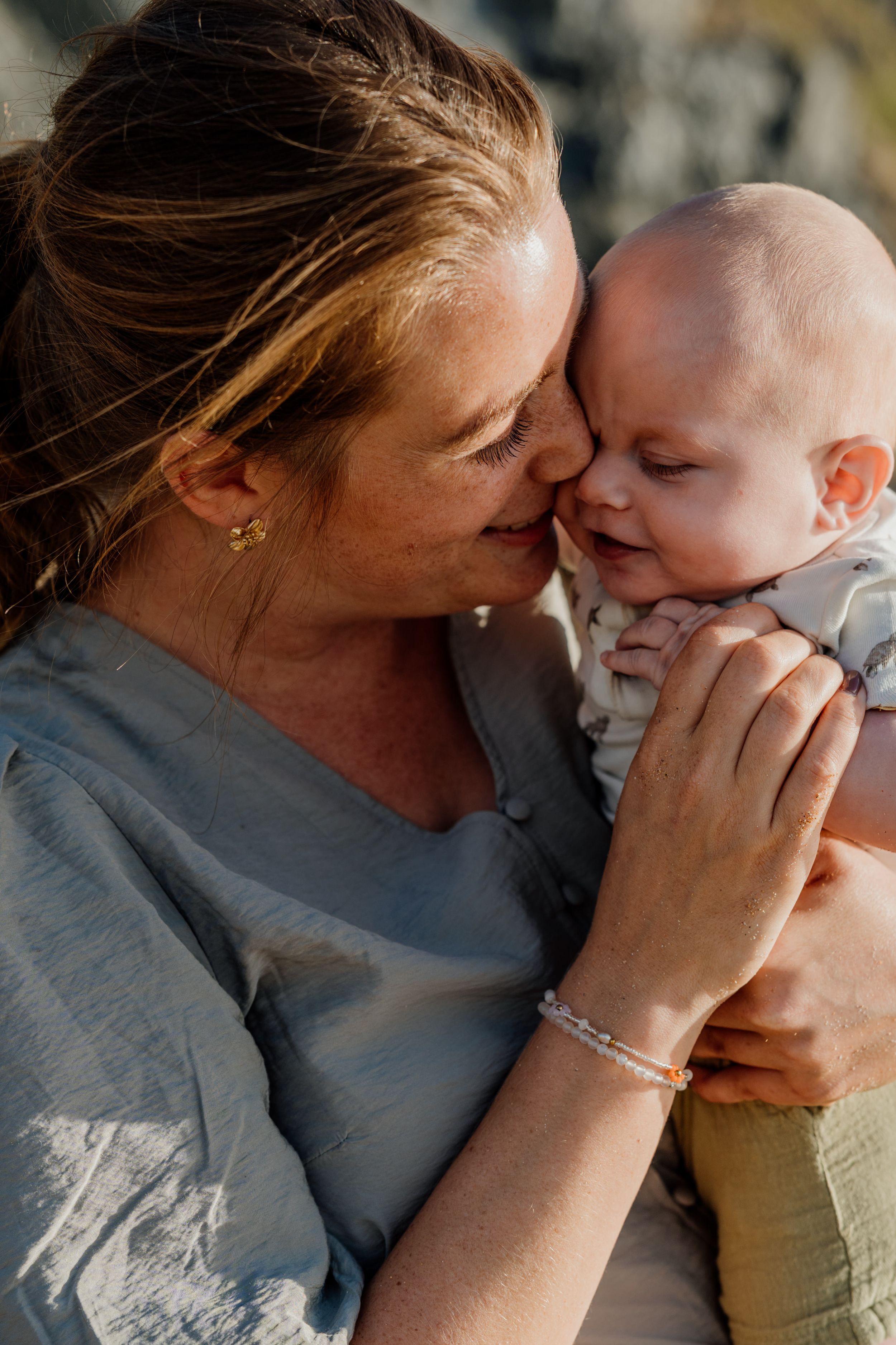 A mother tenderly nuzzles her smiling baby in warm golden sunlight, wearing a blue linen top and beaded bracelet.