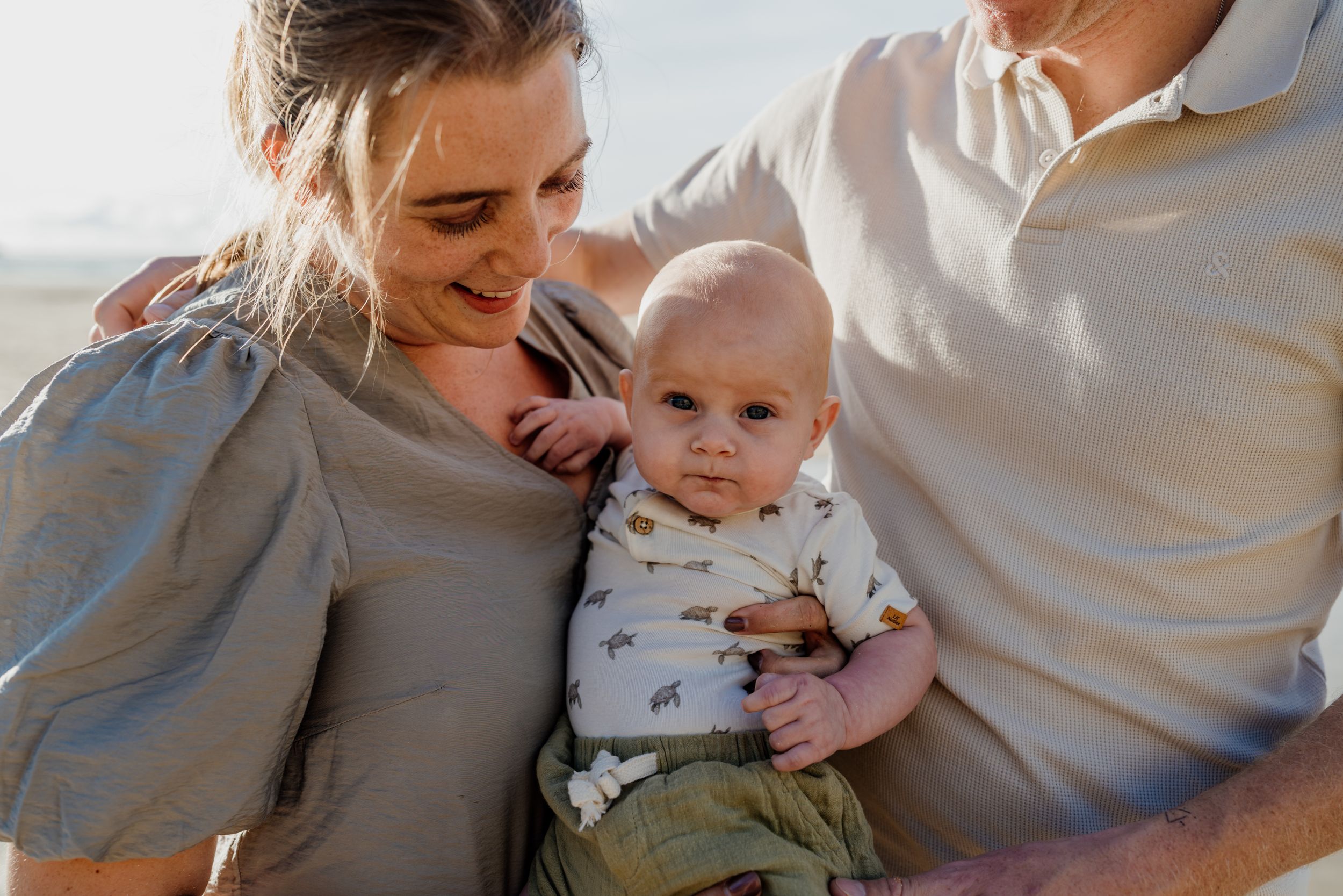 A young couple sitting closely together outdoors, lovingly holding their baby in a warm, natural light setting.