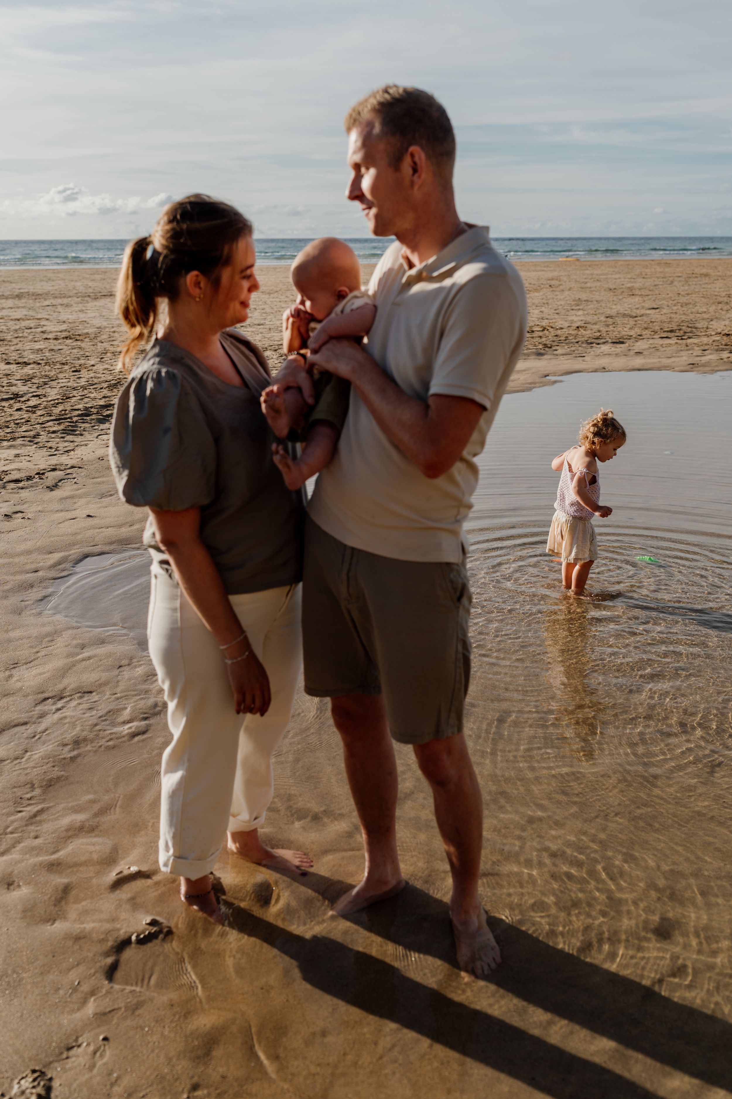 Family with baby stands on sandy beach at sunset, while child plays in shallow ocean water behind them.