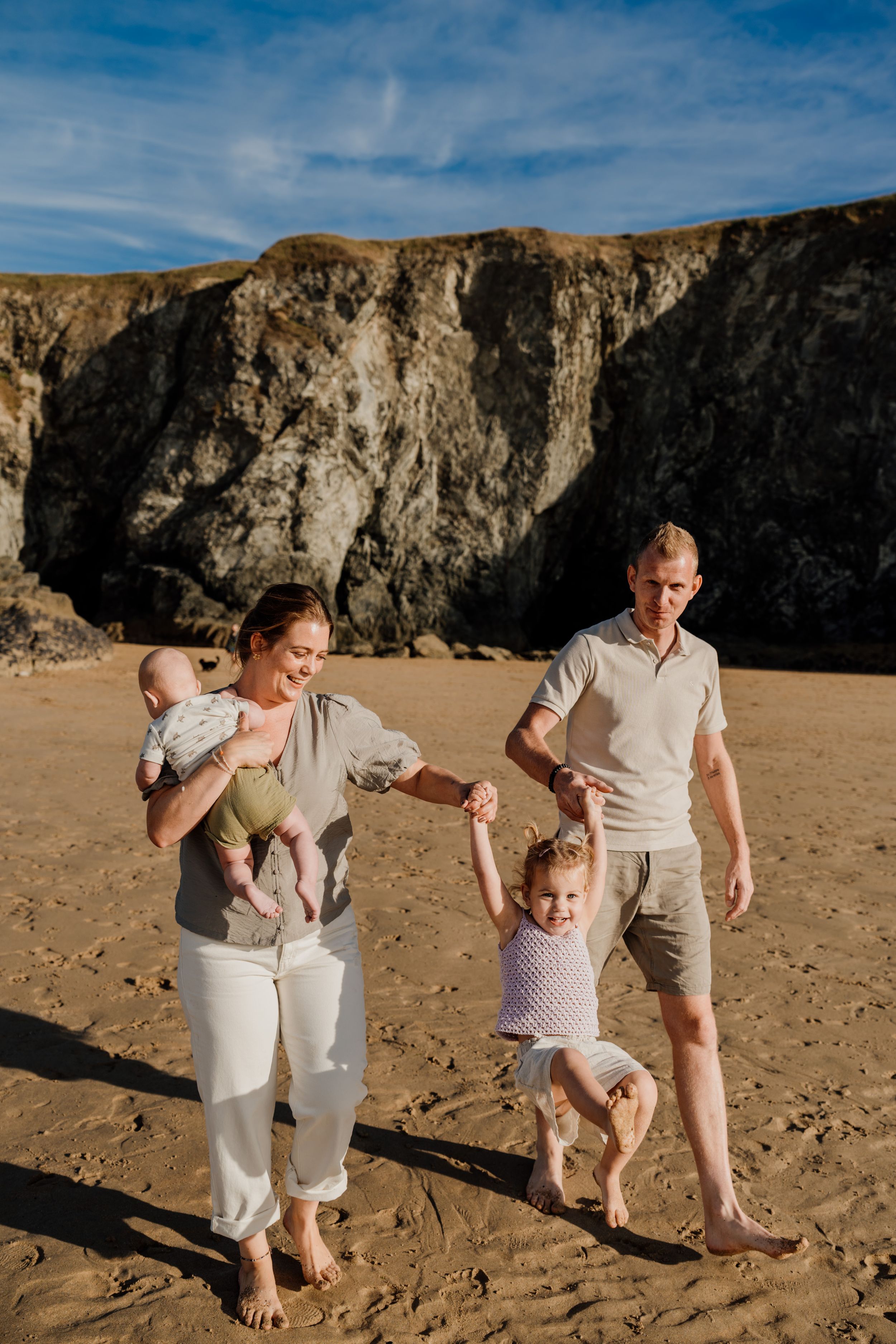 Family of four enjoying a sunny beach day, parents swinging toddler while mother holds baby near rocky cliffs.