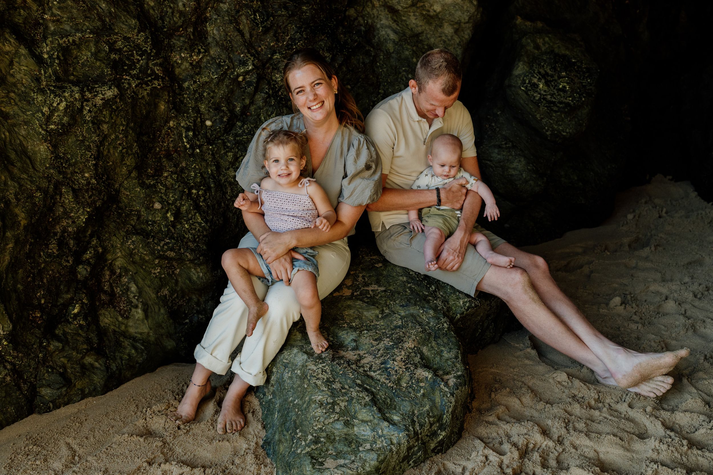 Family of four posing on rocks in a cave, two parents holding young children, smiling warmly in neutral tones.