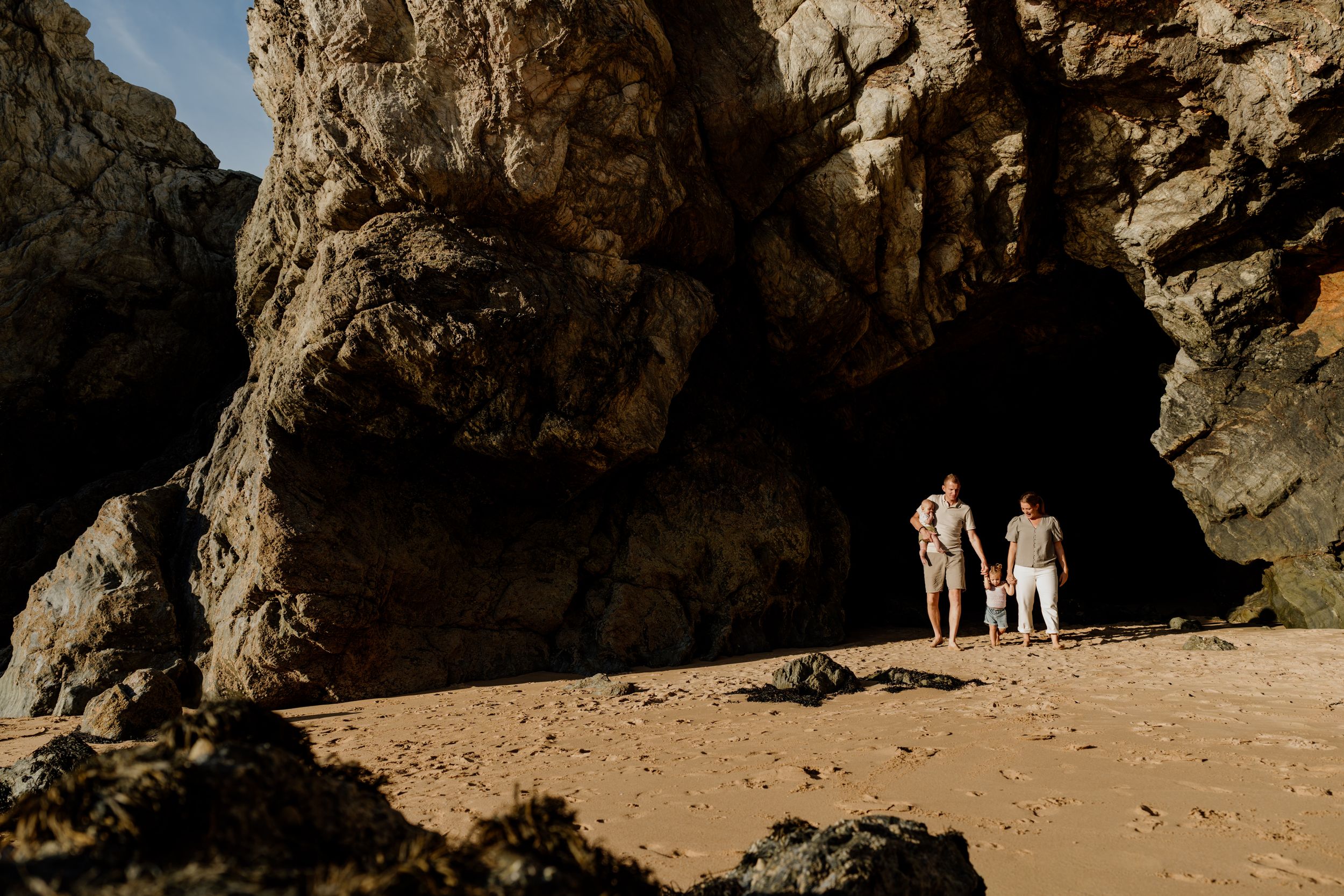 Two people stand on a sandy beach beneath a dramatic natural rock arch cave formation.