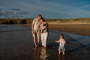 Family of four enjoying a sunset beach walk, parents holding baby while toddler plays in shallow ocean waves.
