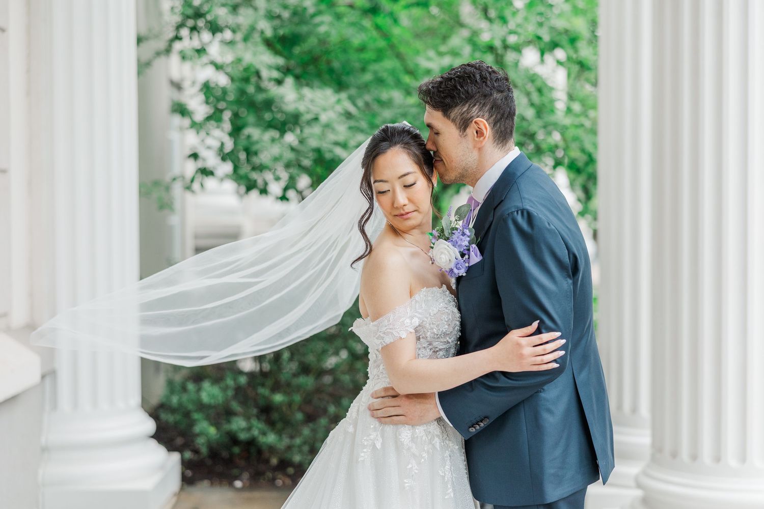 Bride and groom embrace near white columns, veil flowing in wind, lush greenery backdrop.