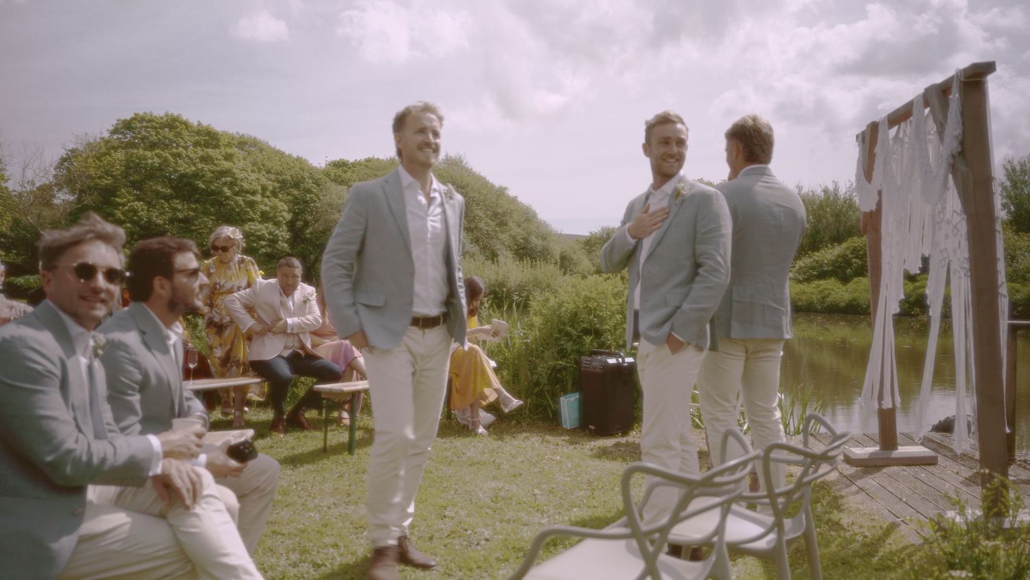 Men in light blue suits gathered at an outdoor wedding ceremony by a pond surrounded by lush greenery.