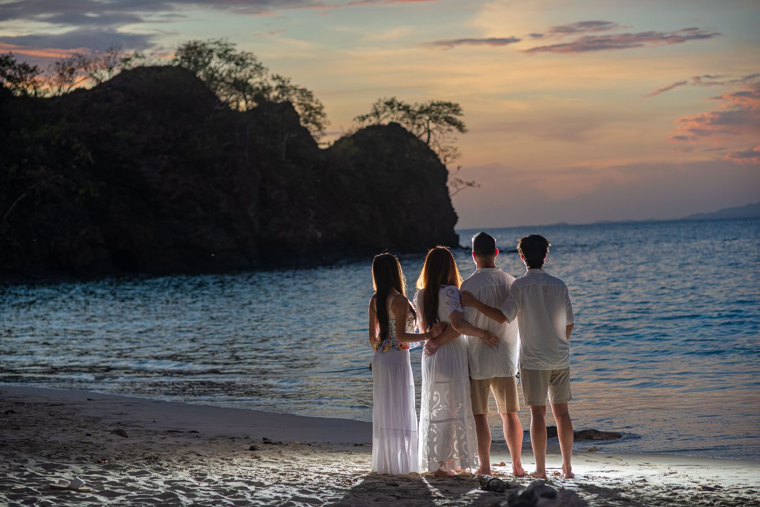 Four people in white attire stand together on a beach at sunset, facing a rocky ocean cove.