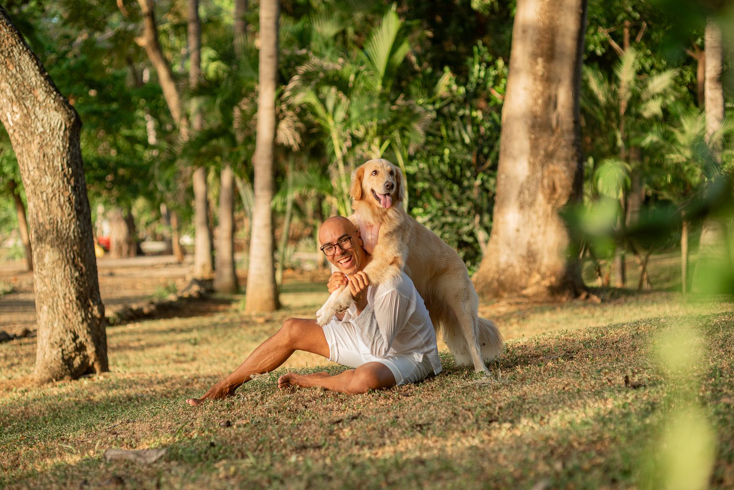 Man sitting on grass in tropical park with golden retriever dog leaning on his shoulders among palm trees.
